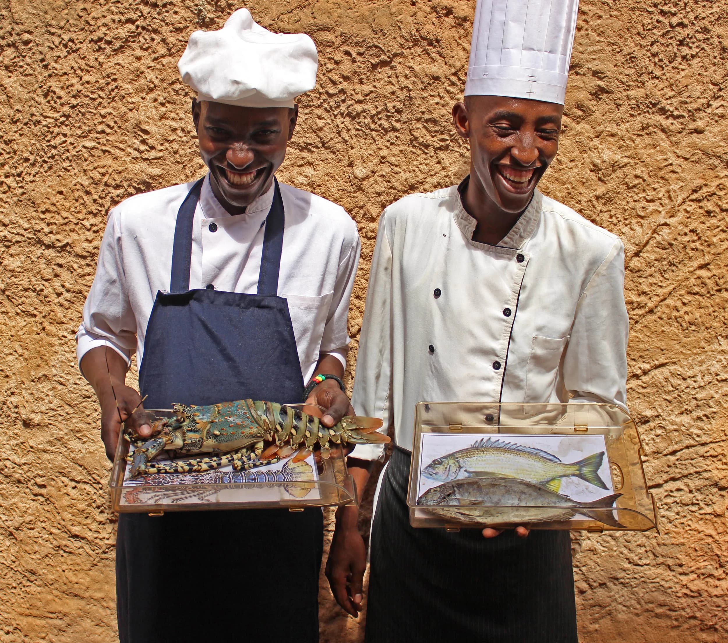 Two laughing chefs holding trays with a fresh spiny lobster and fish from the local catch